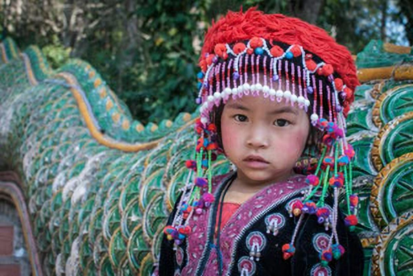 A young girl in traditional vibrant hill tribe clothing and red headdress, representing the cultural diversity of North Thailand.
