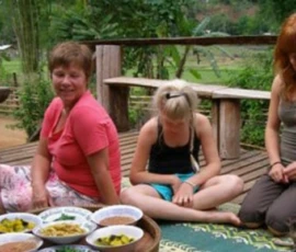 A group of international travelers sitting on a wooden porch with a local host, enjoying traditional snacks.