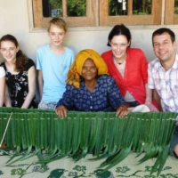 Generations meet over traditional leaf weaving – a hands-on community experience that lies at the heart of Nutty's Andaman Community Explorer tour. Western family with local elderly woman learning traditional leaf weaving community homestay southern Thailand