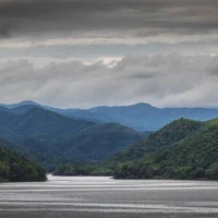 The still waters of Kaeng Krachan Dam reflect the surrounding hills at dusk – a tranquil finale to Day 1 before settling into the Baan Tum Sue homestay. Kaeng Krachan Dam reservoir misty green mountain landscape Phetchaburi Thailand